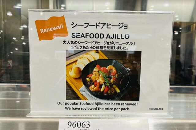 コストコ倉庫店の紹介チラシ。『NEW』タグ付きで大きくシーフードアヒージョの写真を配置。エビ・イカ・ホタテがガーリックオイルに絡む温かいイメージ、黒地に白文字で商品名『SEAFOOD AJILLO』と商品番号『ITEM#96063』、おすすめの食べ方『バゲットやワインと一緒に』などのキャッチコピーが掲載された販促ポスター風デザイン
