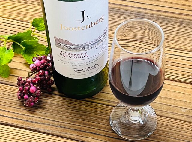 コストコ「ユステンバーグ カベルネ・ソーヴィニヨン」ITEM#518726。赤ワインをグラスに注いだシーンとボトルを並べた写真。深みのあるルビーレッドの色合いと芳醇なカシスやプラムの香りが漂う、南アフリカ・ステレンボッシュ産のフルボディ赤ワイン。
