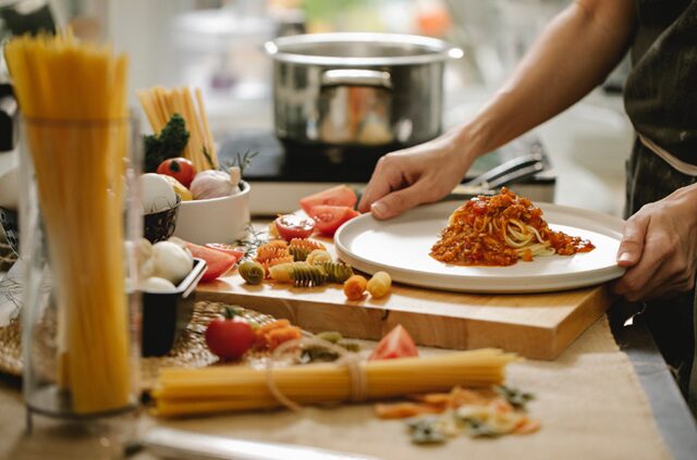 キッチンで赤ワインに合うトマトソースパスタを盛り付けている様子。湯気が立つパスタに濃厚なトマトソースを絡め、仕上げにバジルとチーズをトッピング。奥にはグラスに注がれた赤ワインが見える家庭的で温かいシーン。